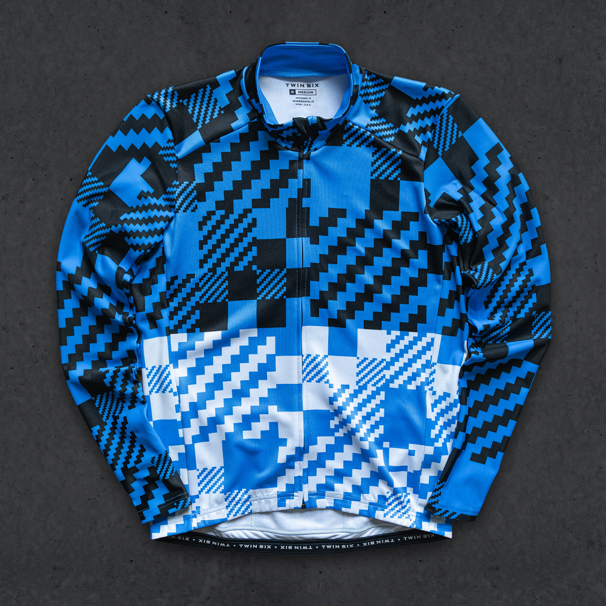 A long-sleeve thermal jersey with a bold geometric pixelated pattern in blue, black, and white, laid flat on a dark textured background.