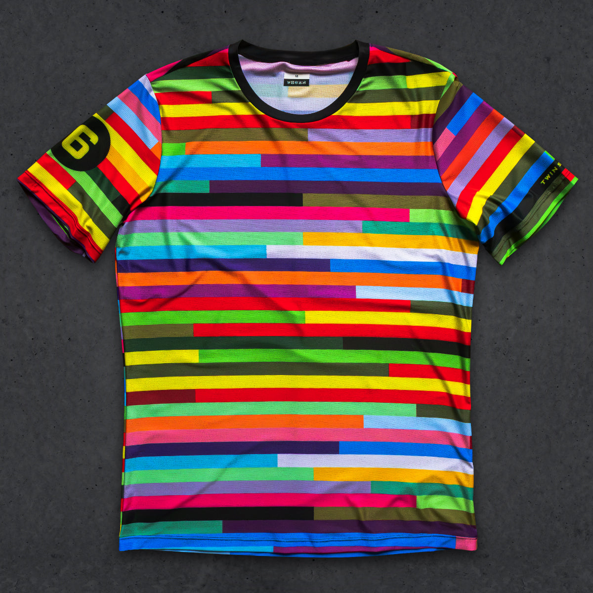 A short-sleeve crewneck t-shirt featuring a vibrant, multicolored horizontal stripe pattern with varying widths, laid flat on a dark textured background. The left sleeve features a black circle with a yellow stylized 'G' logo, and the inner collar label shows the brand 'G-DRIVE'.