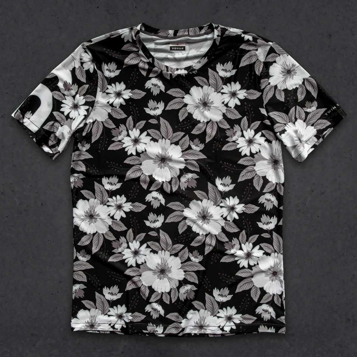 A short-sleeve crewneck t-shirt with an all-over black, white, and gray floral print, laid flat on a dark textured background.