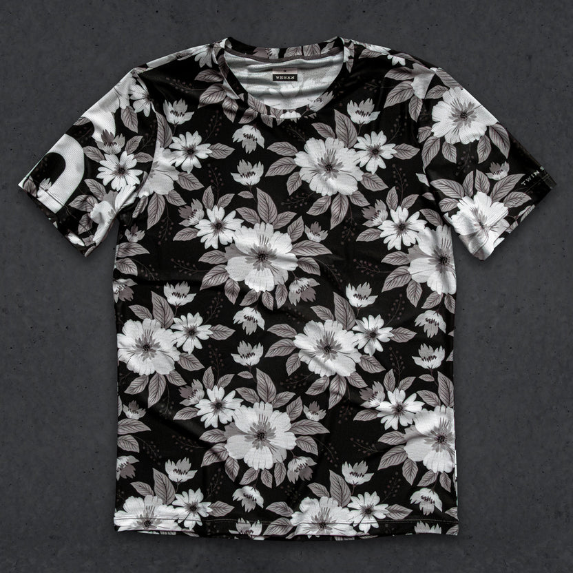 A short-sleeve crewneck t-shirt with an all-over black, white, and gray floral print, laid flat on a dark textured background.